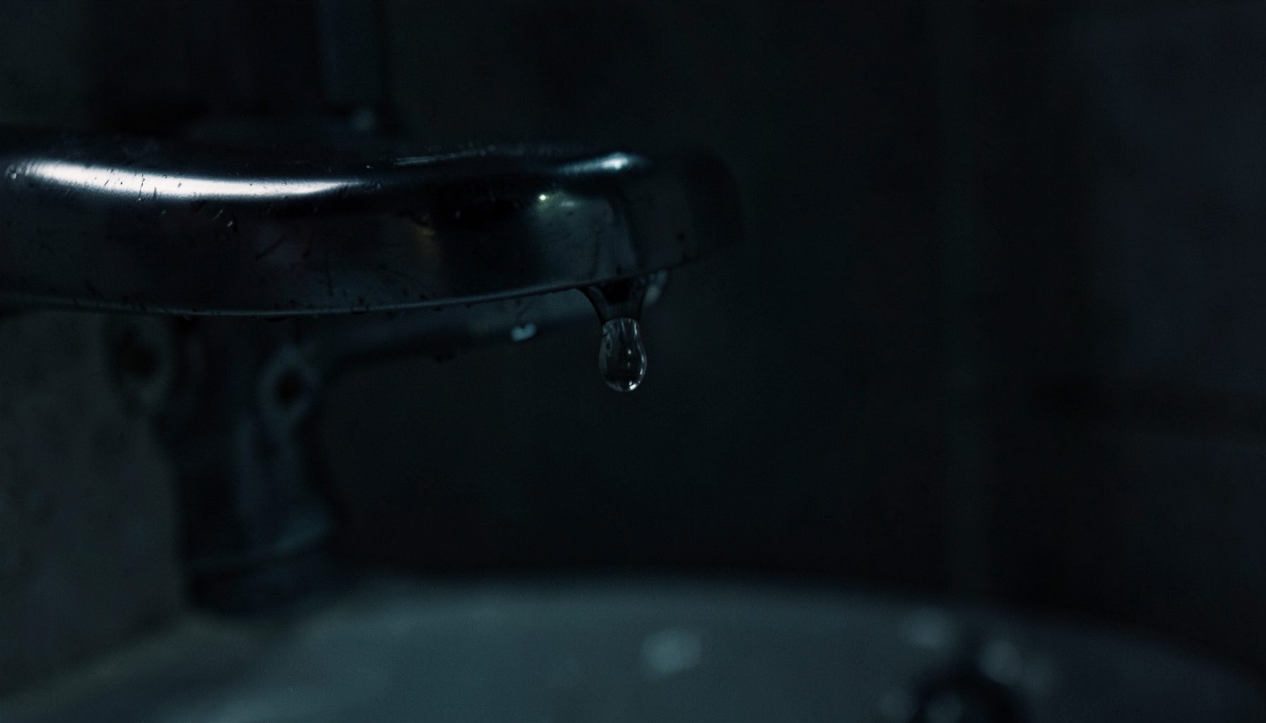 Robinet de salle de bains vu de près, goutte d'eau suspendue, éclairage faible et tons métalliques froids.