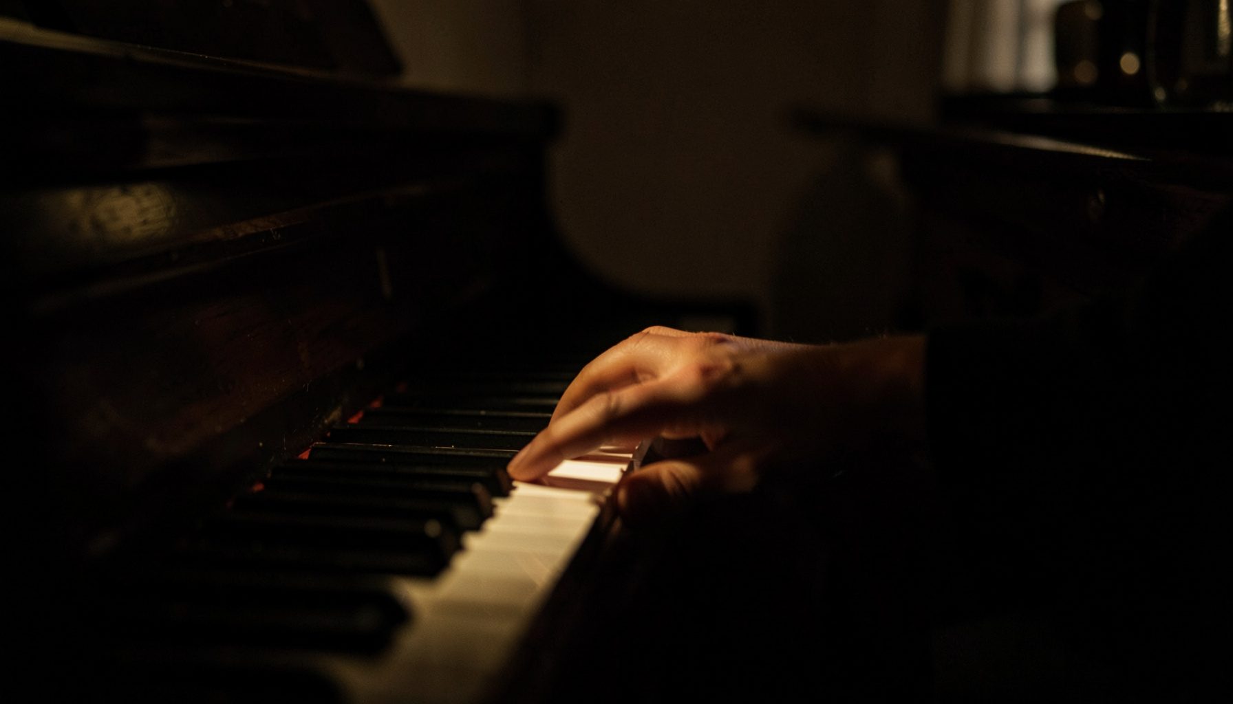 Photographie sombre d'un intérieur chaleureux, une main posée près d'un clavier de piano éclairé par une faible lumière ambrée, ambiance intime et recueillie.